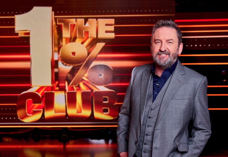 Lee Mack en un traje, parado frente al "El club del 1%" logo.