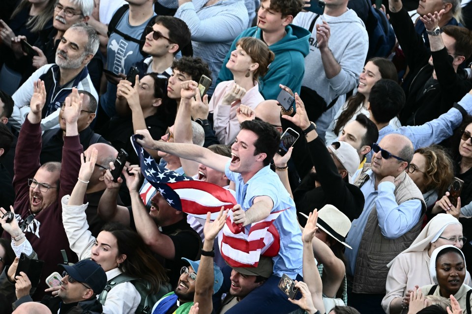 Una multitud de personas que animan, sosteniendo una bandera estadounidense, reaccionando al anuncio de un nuevo papa.