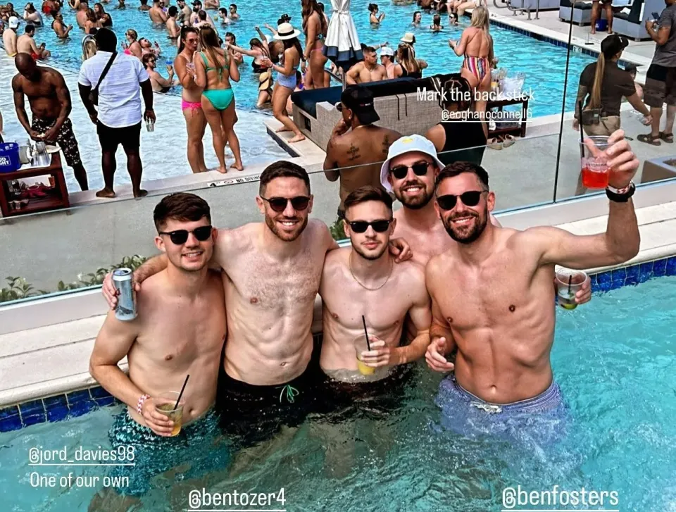 Cuatro hombres sin camisa en una piscina con bebidas.