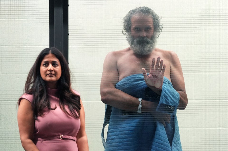 Un hombre y una mujer vistos a través de una partición de malla de alambre; El hombre no tiene camisa y sostiene una mano.