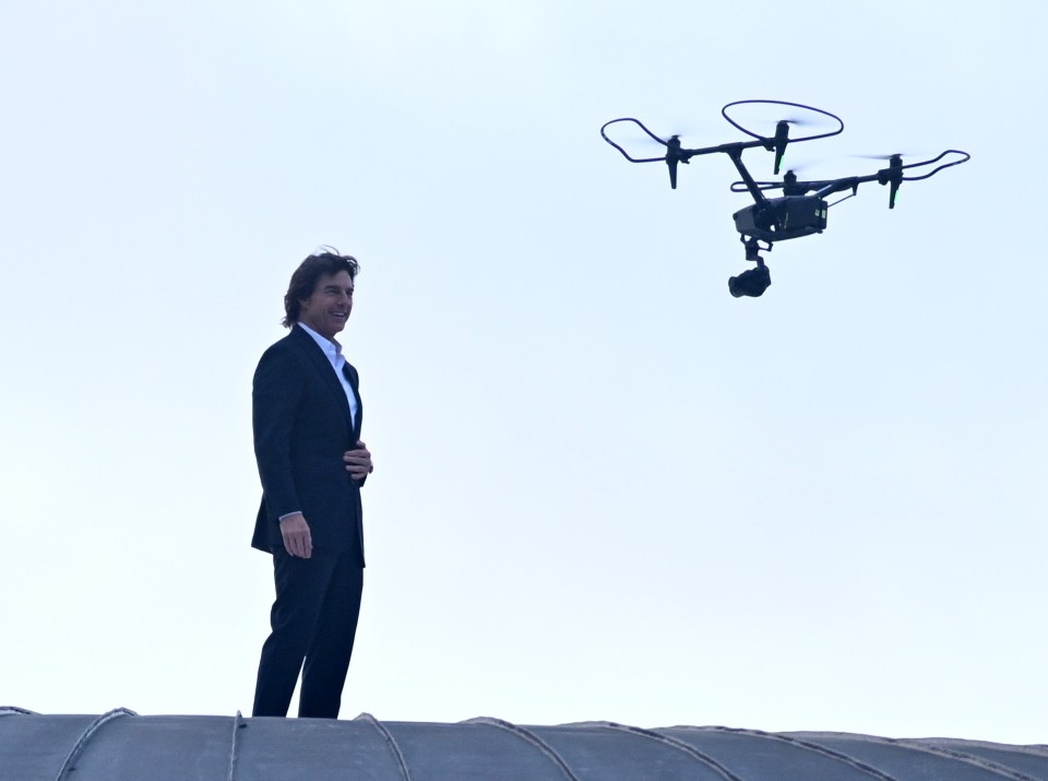 Tom Cruise parado en una azotea con un dron en lo alto.