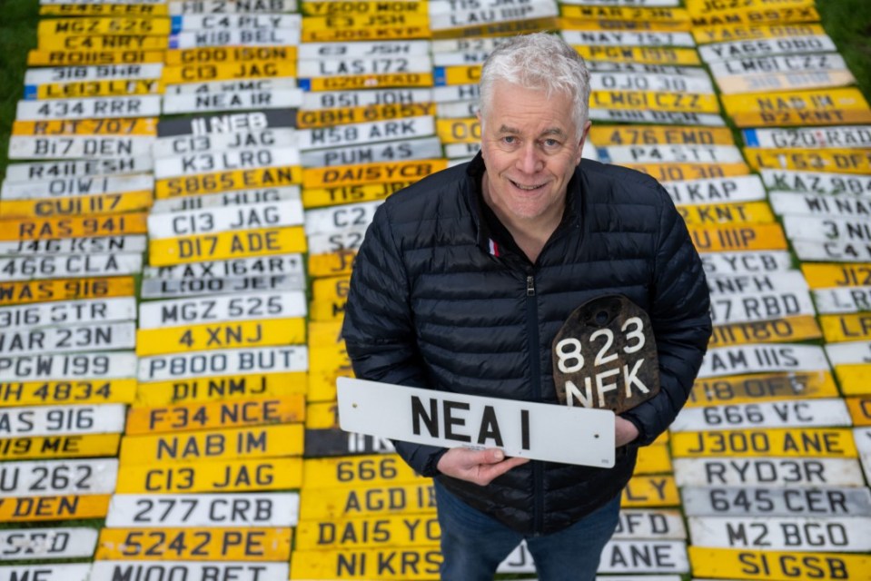 Hombre sosteniendo una matrícula, rodeado por cientos de otras placas de números.