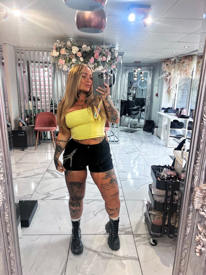 Una mujer tatuada en una blusa amarilla y pantalones cortos negros se toma una selfie en una peluquería.