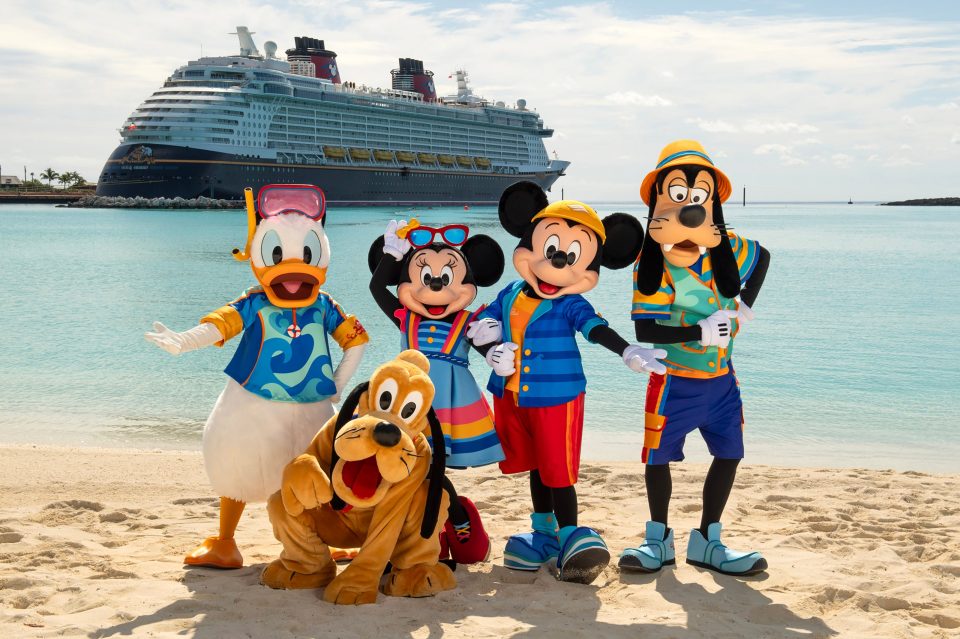 Mickey Mouse, Minnie Mouse, Goofy y Donald se agachan en la vestimenta de la playa en una playa con un crucero de Disney en el fondo.