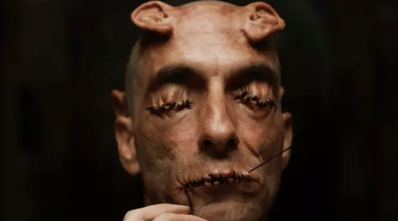 Primer plano de un hombre con ojos y boca cosidos, y pequeños cuernos en la cabeza.