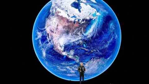 Un hombre se para en un escenario que da una charla frente a una imagen de pantalla gigante de un planeta azul tierra rodeada por la oscuridad del espacio