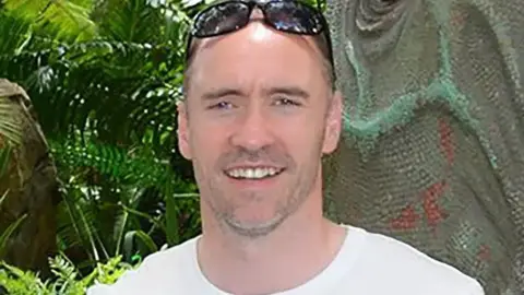 Paul Doyle tiene el cabello corto y oscuro con gafas de sol en la cabeza y está vistiendo una camiseta blanca.