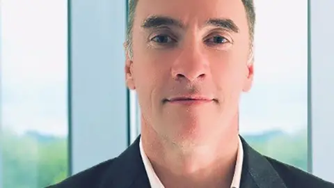 Unknown Paul Doyle, quien tiene el cabello gris peinado en un quiff, sonríe a la cámara mientras usa un saco negro y una camisa blanca sin corbata