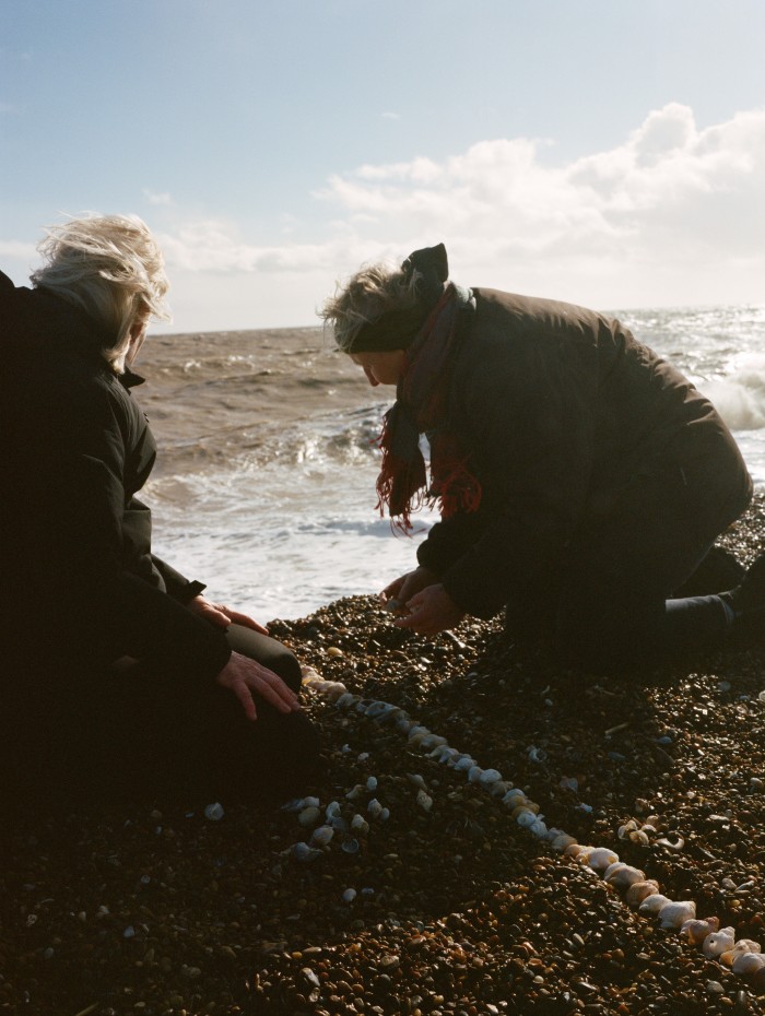 Deux femmes agenouillées sur une plage de galets disposent des coquillages en alignement avec la mer en arrière-plan