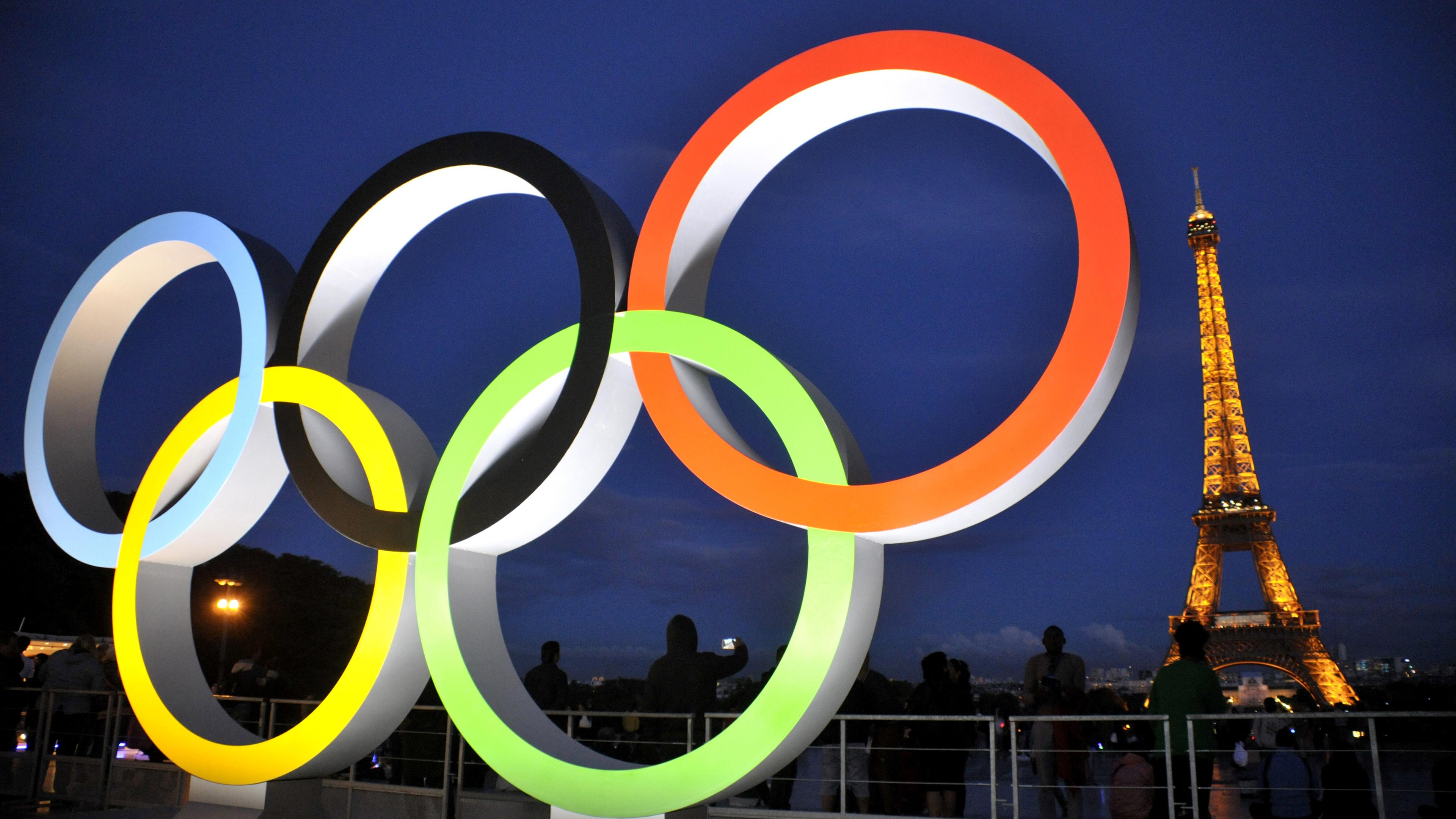 Les anneaux olympiques illuminés au premier plan.  La Tour Eiffel illuminée en arrière-plan.