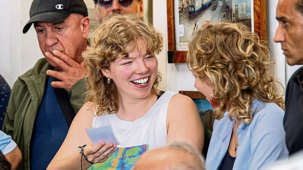 Une jeune femme aux boucles blondes se moque d'une autre jeune femme, tenant dans sa main une carte d'enchère provenant d'une vente aux enchères