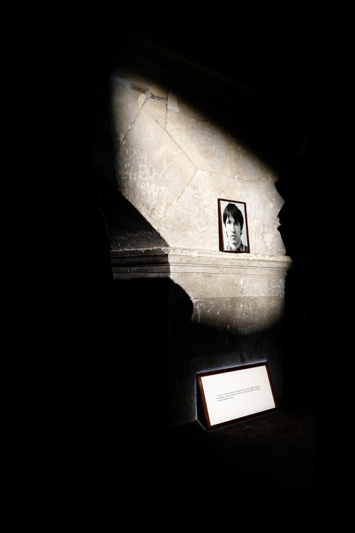 Une petite photographie en noir et blanc du visage d'un homme accroché à un mur de pierre, éclairé par un faisceau de lumière vive