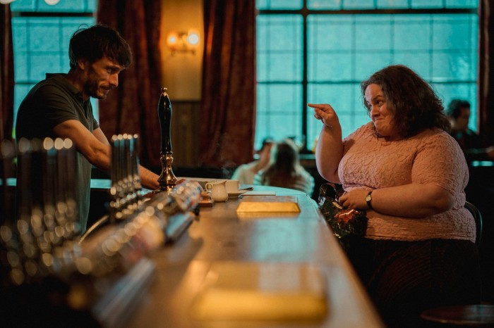 Une femme portant un haut rose pointe du doigt un barman qui a l'air dubitatif