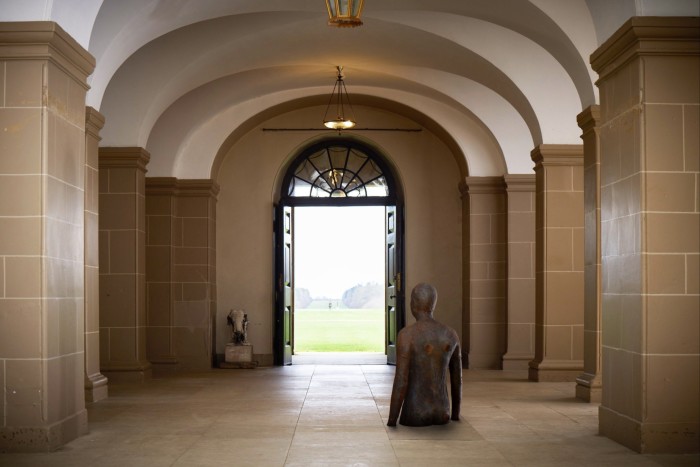 Une statue d'une personne jusqu'à la taille dans un couloir avec des arches qui donne à travers un cadre de porte sur un champ herbeux avec une autre statue au loin