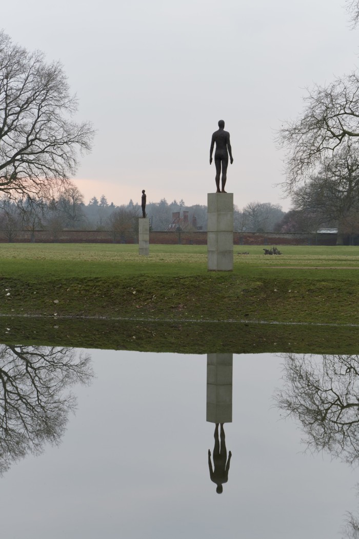 Une grande statue en fonte d'une personne sur un socle se reflétant dans un bassin d'eau. Il y a des arbres de chaque côté