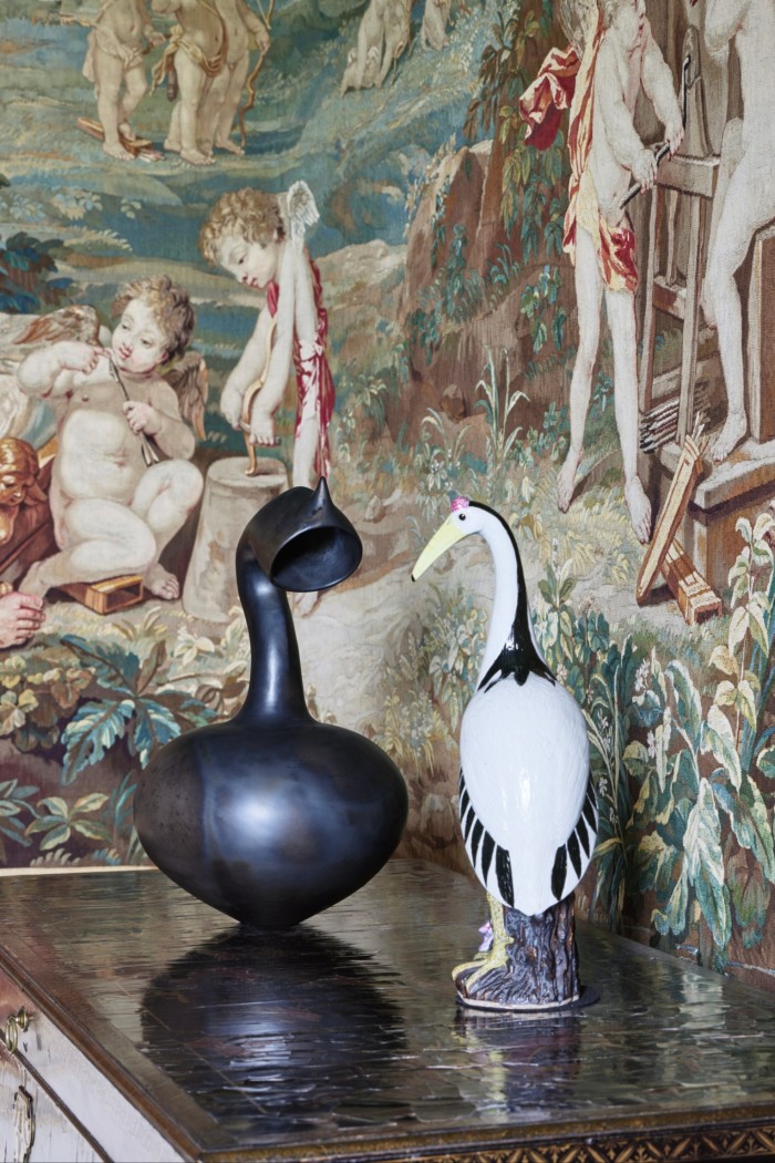 Deux sculptures posées sur une table devant une vieille tapisserie. L'une des sculptures est une figure en céramique détaillée d'un oiseau blanc, l'autre est un vase sombre à long col