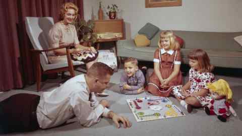 une jeune famille sur le sol jouant à un jeu de société pendant que maman fait du crochet sur une chaise, 1962