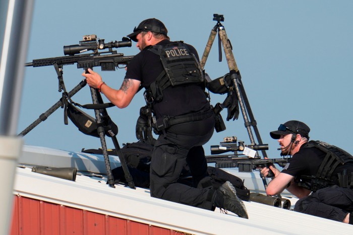 Deux hommes vêtus de noir sont assis sur un toit blanc. L'un est à genoux, tenant un grand fusil noir sur un trépied et regardant à travers sa lunette vers la gauche. Le deuxième est allongé sur le ventre et regarde également à travers la lunette de son fusil.