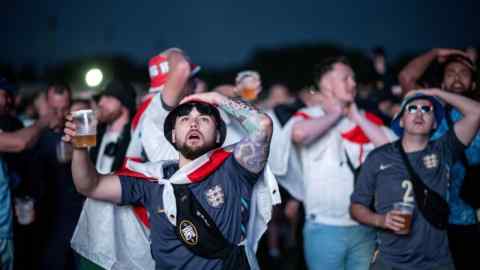 Des jeunes hommes en maillot de football bleu ou blanc, certains drapés dans le drapeau anglais, lèvent les yeux d'un air tendu. Certains d'entre eux se tiennent la tête avec une main.