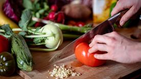 Une personne coupe une tomate sur une planche à découper. Différents types de légumes se trouvent à côté.