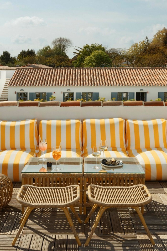 Sur une terrasse ensoleillée, avec un bâtiment blanc et bas derrière, des tabourets en osier et un banc avec des coussins rayés blancs et jaunes. Des boissons et une assiette d'huîtres sont posées sur la petite table au milieu