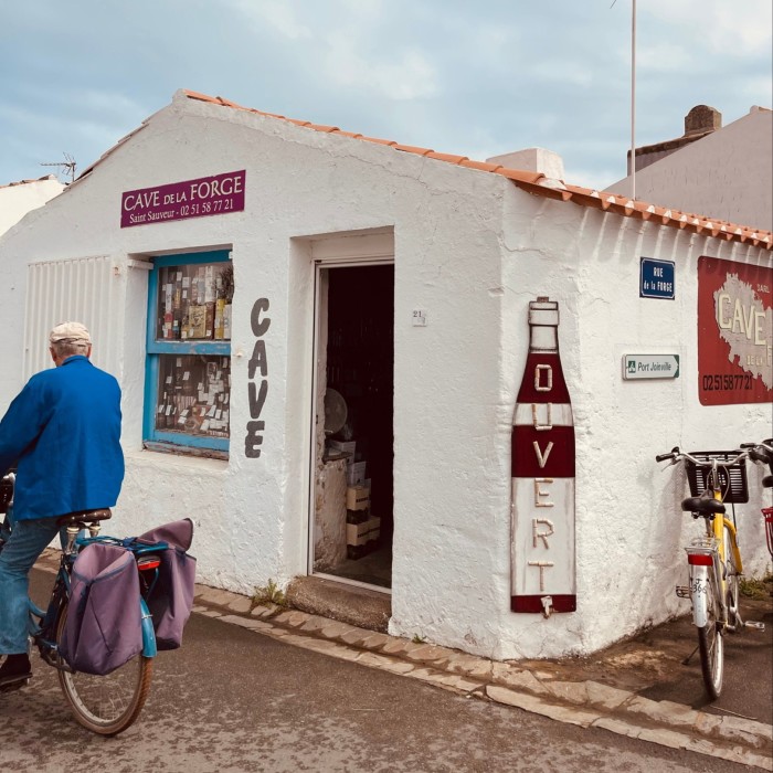 Un homme portant une veste bleue passe à vélo devant un magasin blanchi à la chaux avec une grande pancarte en forme de bouteille sur laquelle est écrit « ouvert » 