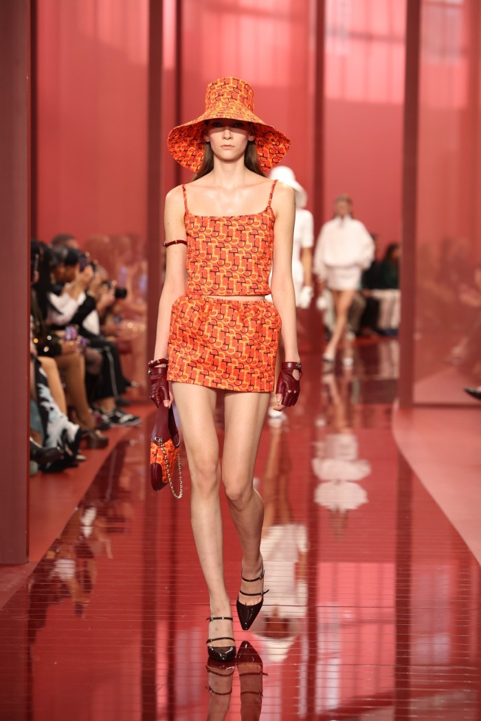 Un mannequin féminin sur un podium rouge portant une mini-jupe orange vif et un chapeau à large bord de couleur similaire