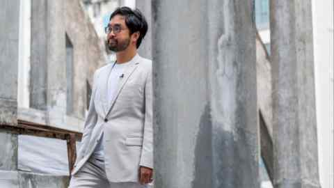 Adrian Cheng se tient devant une structure en béton du Théâtre d'État de Hong Kong. Il porte un costume clair et des lunettes.