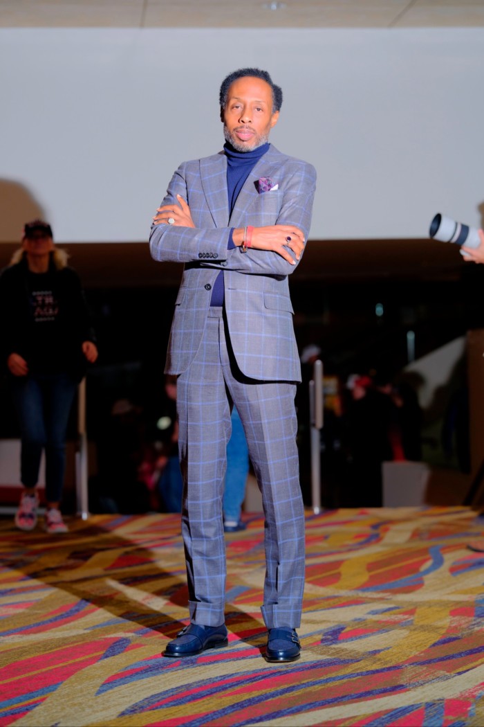 Dwayne Hardin se tient debout, les bras croisés, vêtu d'un costume à carreaux gris et d'un col roulé foncé.