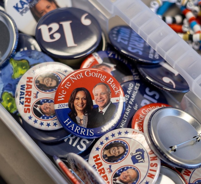 L'image montre une collection de boutons de campagne politique pour Harris et Walz à l'intérieur d'un récipient en plastique.