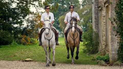 Deux hommes portant des lunettes de soleil, des chemises blanches, des bottes d'équitation et des jodhpurs beiges chevauchent des chevaux adjacents devant un bâtiment en pierre dans une campagne idyllique