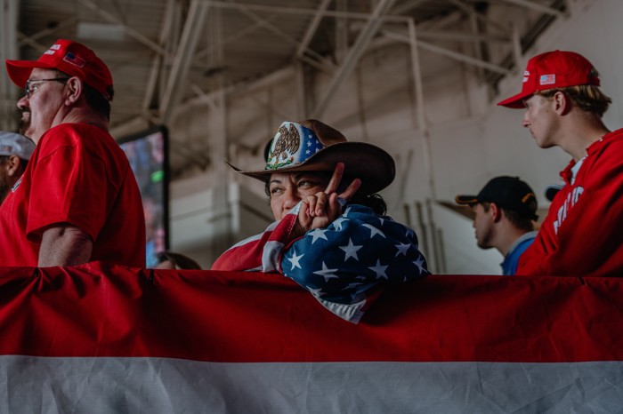 Une femme est assise, enveloppée dans un drapeau étoilé. Elle porte un chapeau de style cowboy qui montre une partie des drapeaux mexicain et américain. À côté d'elle se tient un homme en chemise rouge et casquette de baseball rouge.