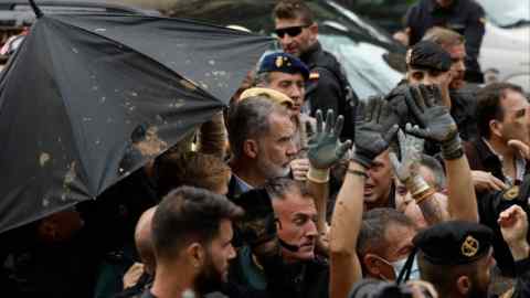 Un homme à la barbe grise, au milieu d'un groupe d'autres hommes, certains en uniforme de police, est en partie abrité par un parapluie éclaboussé de boue.