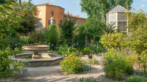 Un jardin avec une haute serre hexagonale qui fait écho à la forme du bâtiment situé derrière. Au premier plan se trouve une fontaine circulaire en pierre