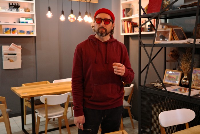 Un homme portant un pull rouge, un bonnet rouge assorti et des lunettes teintées de rouge se tient dans un café. L'arrière-plan présente des tables, des chaises, des étagères et des décorations en bois, ainsi qu'un éclairage chaleureux.