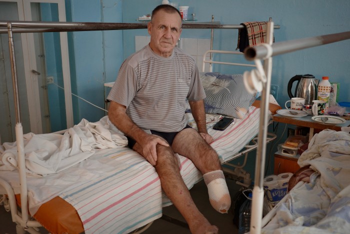 Un homme au pied bandé est assis sur un lit d’hôpital. Il porte une chemise rayée et un short, avec une expression calme. Le lit est entouré d'objets personnels, notamment une table avec de la nourriture, des boissons et des fournitures médicales.