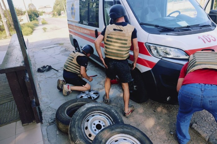 Trois individus portant des casques de protection et des gilets pare-balles réparent une ambulance endommagée lors d'un incident à Antonivka, en Ukraine. L'ambulance est garée au bord d'une rue, et plusieurs pneus et outils démontés sont visibles au sol à proximité