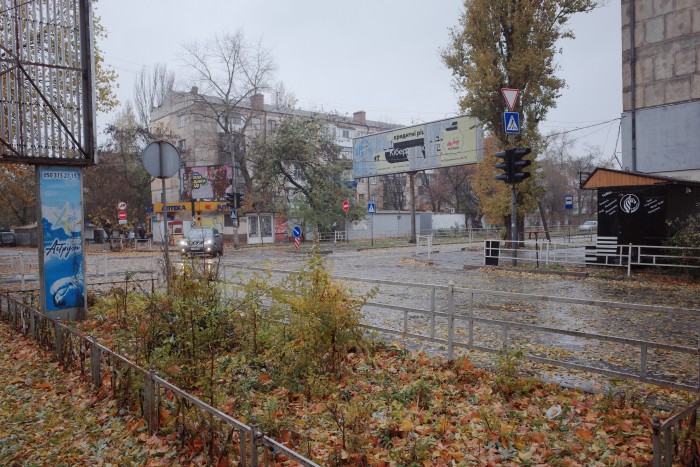 Une intersection de rue pluvieuse à Kherson montrant des feuilles d'automne tombées éparpillées sur le sol et une chaussée mouillée. La scène comprend des feux de circulation, des panneaux de signalisation et des bâtiments commerciaux avec des panneaux d'affichage. Une voiture avec les phares allumés traverse l'intersection et la zone est entourée d'une végétation clairsemée et d'infrastructures urbaines.