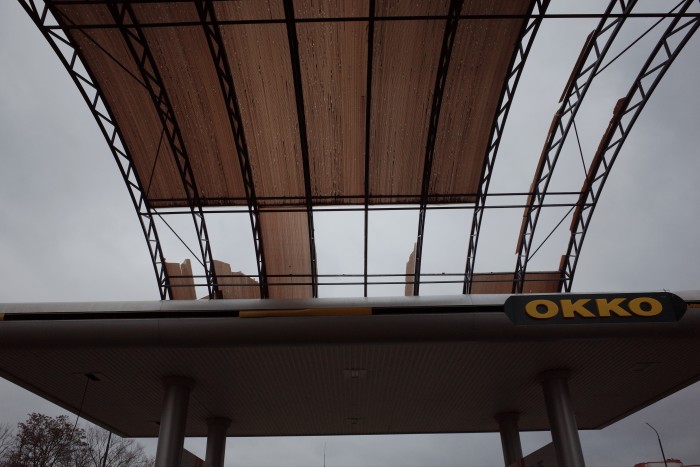 Le toit endommagé de la station-service Okko, avec de grandes sections de la verrière manquantes ou cassées. La charpente métallique est exposée sur un ciel nuageux. Le panneau de la station-service avec le logo « OKKO » est visible sur le bord de l'auvent.