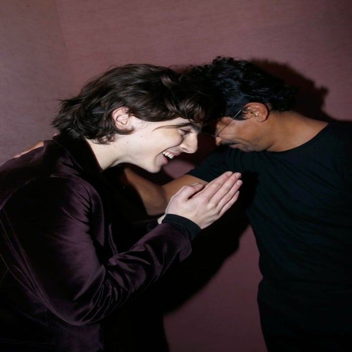Deux personnes partagent un moment joyeux, l'un aux cheveux longs dans une tenue élégante et l'autre habillée avec désinvolture, les deux semblent rire ou sourire