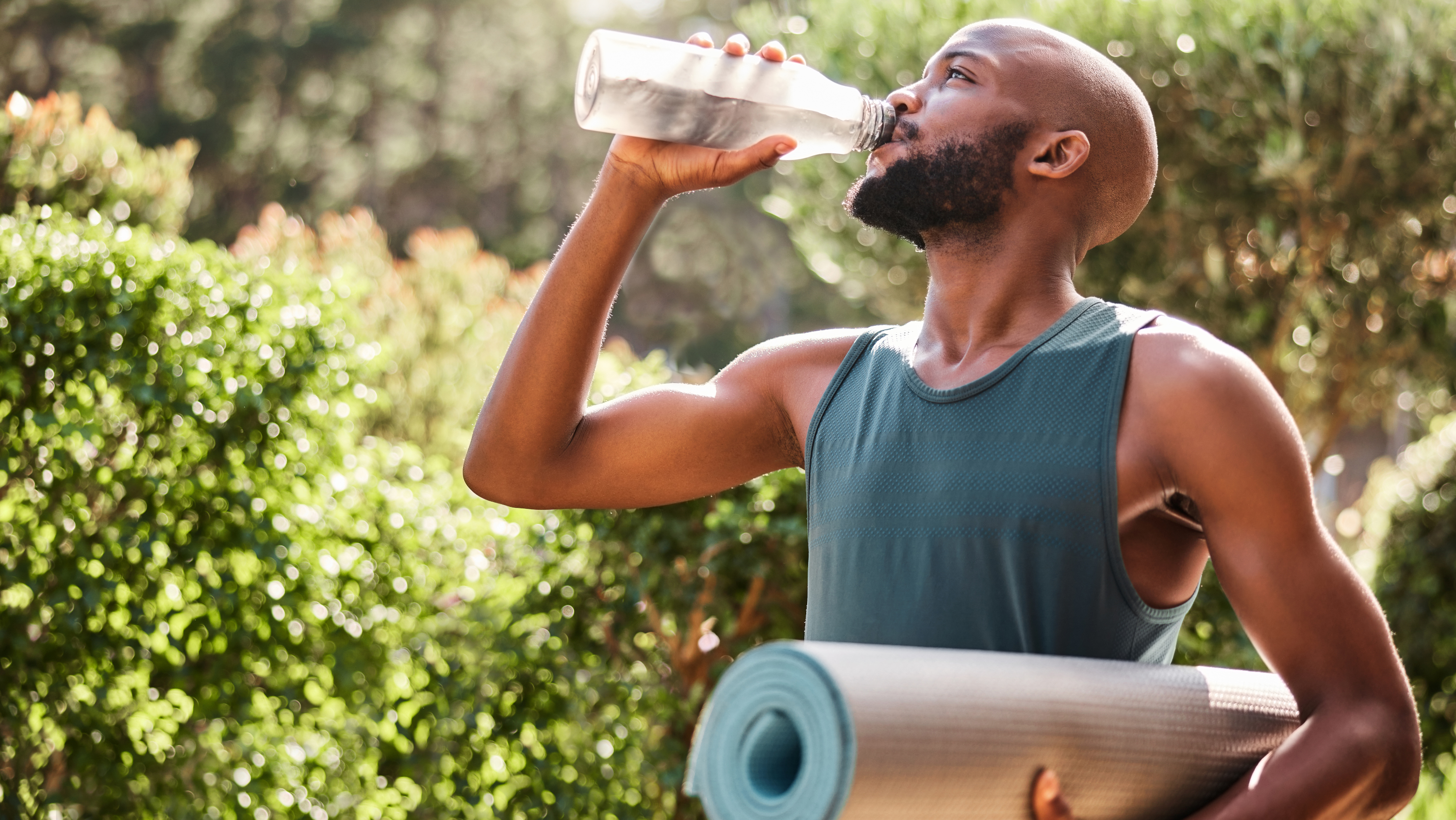 bir elinde yoga matı taşıyan, diğer elinde ise su şişesinden su içen bir adamın fotoğrafı