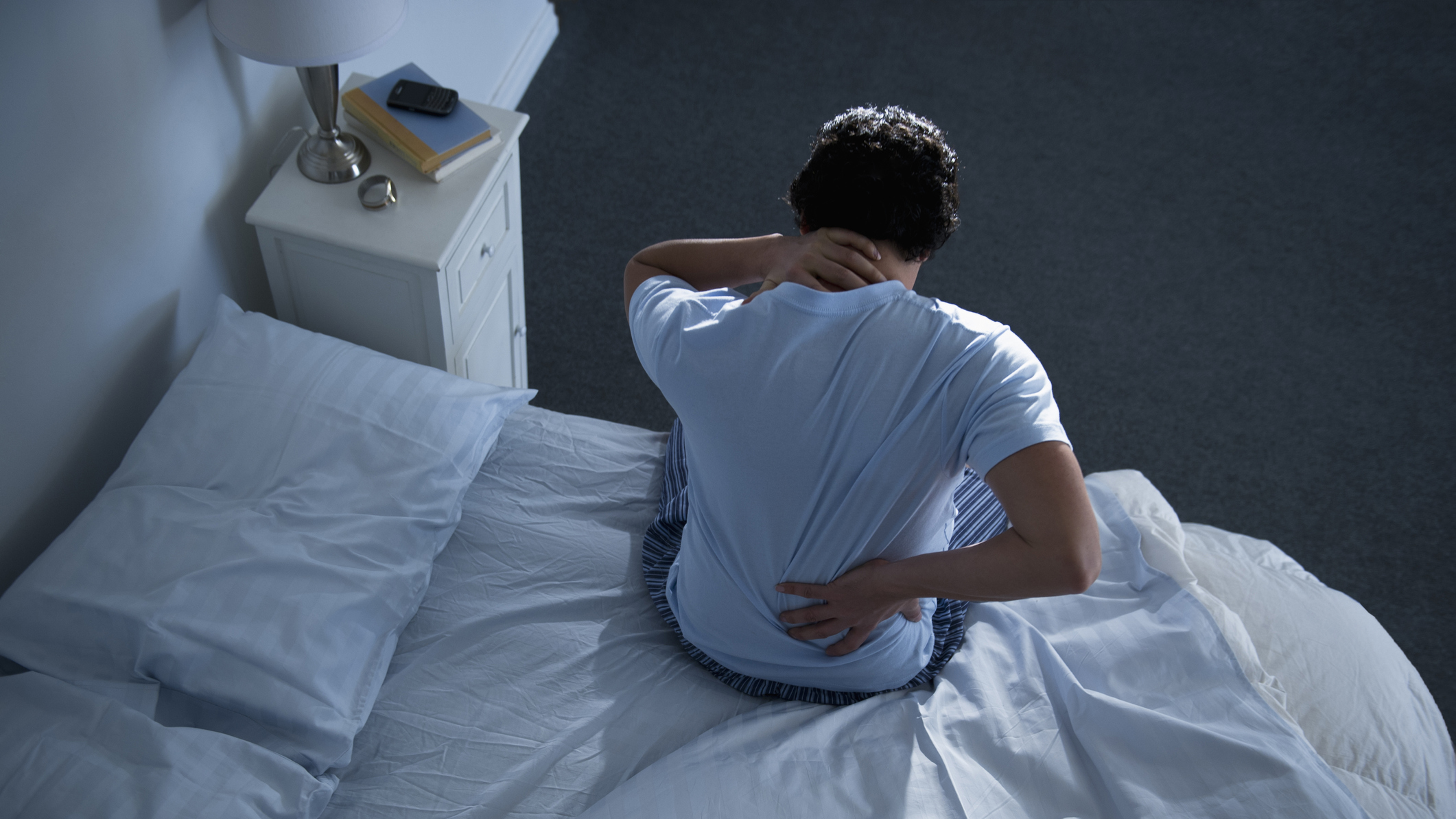 Bir adam geceleri sırt ağrısı yaşadığı için bir elini beli üzerinde tutarak yatakta oturuyor