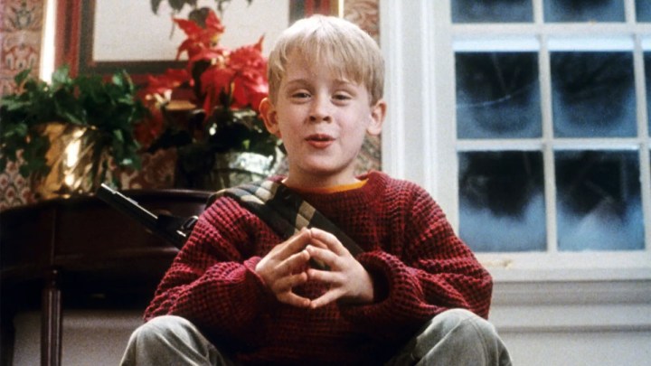 "Evde Tek Başına" filminde genç bir çocuk elinde silahla merdivenlerde oturuyor.