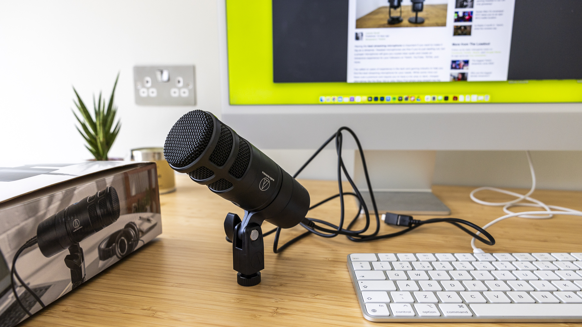 Birisinin masasında, bir kutunun ve klavyenin yanında bulunan mikrofonu gösteren Audio Technica inceleme görseli.