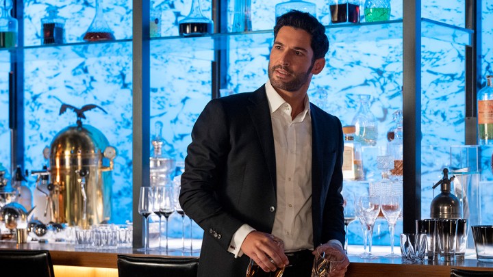 Lucifer Morningstar rolünde Tom Ellis, takım elbiseyle barının yanında duruyor ve Netflix'teki Lucifer filminden bir sahnede birine bakıyor.