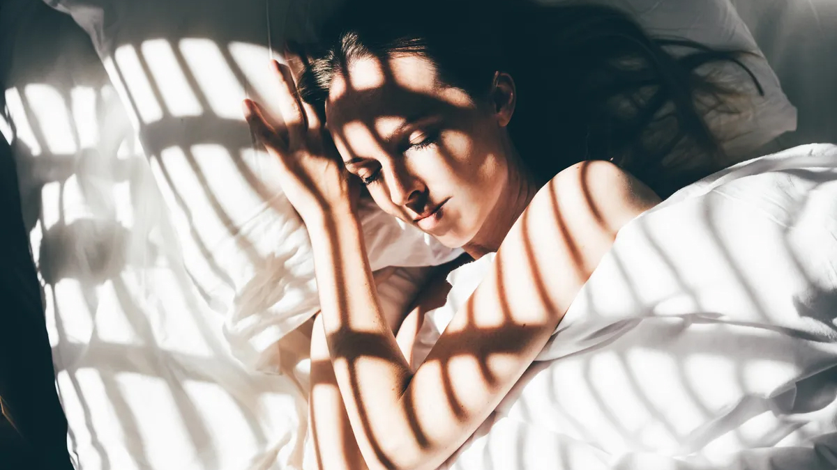 Uzun siyah saçlı bir kadın beyaz bir yorganın altında çıplak kollarıyla uyuyor