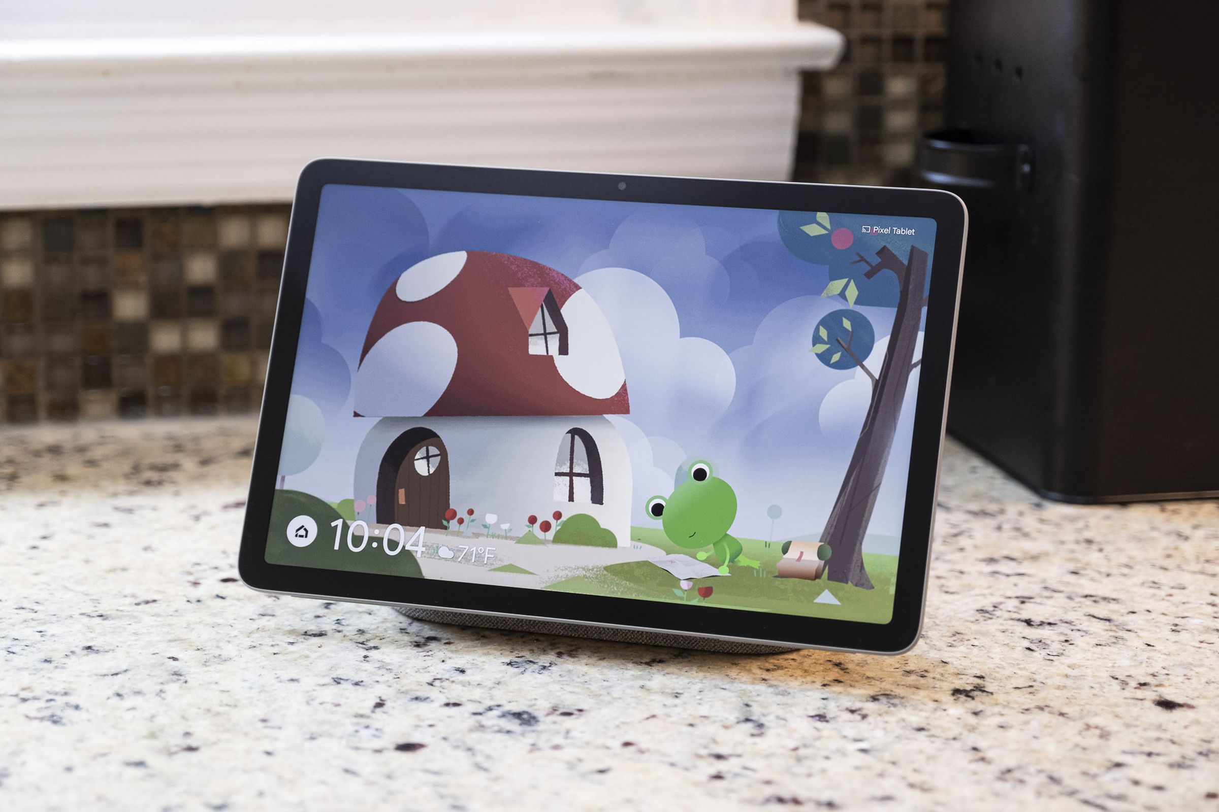 Hoparlör yuvasına monte edilmiş ve Hava Durumu Kurbağası ekran koruyucusunu gösteren bir Piksel Tablet.