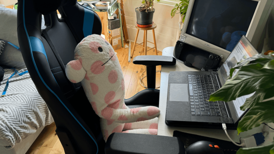 Dizüstü bilgisayar, CRT monitör ve bitkilerle dolu bir masadaki oyun koltuğunda bir peluş oyuncak oturuyordu