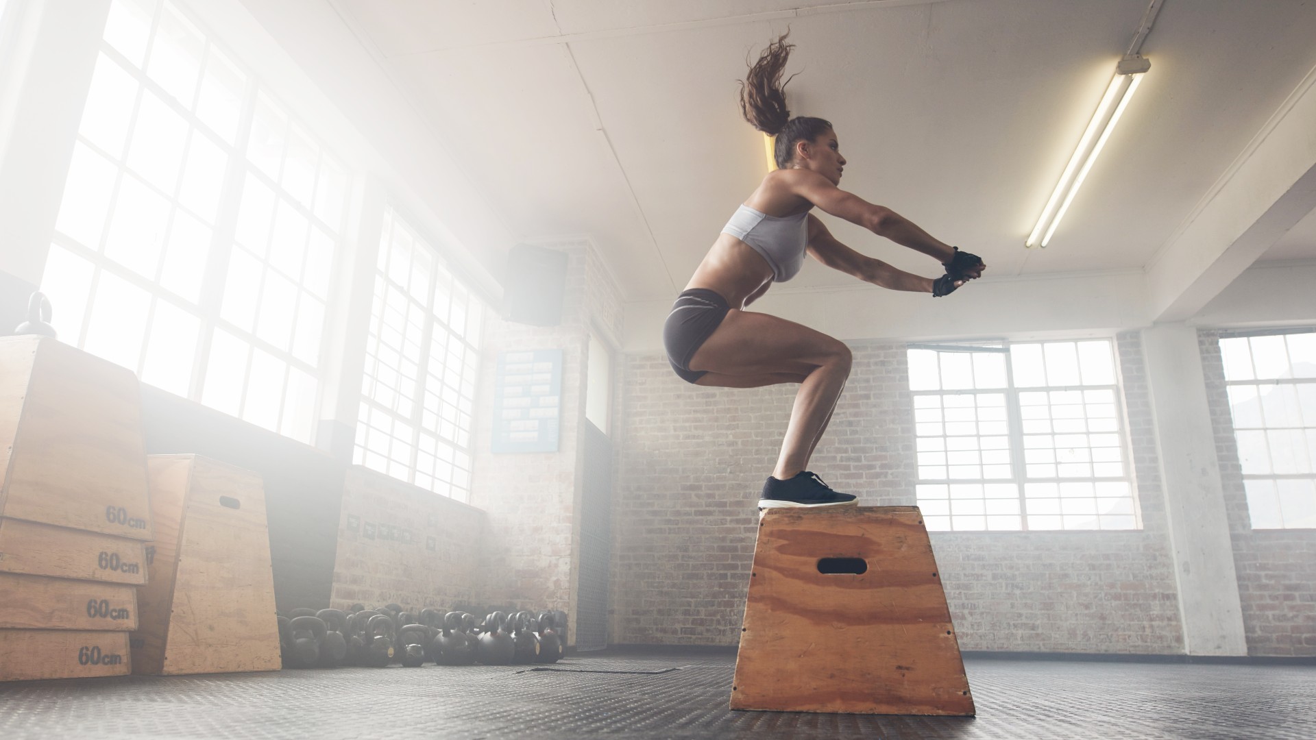 Bir depo spor salonunda kutuya atlayarak vücut ağırlığı antrenmanı yapan kadın, her iki ayağıyla bir kutunun üzerine iniyor