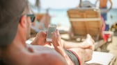 Beysbol şapkası takan bir adam plaj şezlongunda yatıyor ve akıllı telefonuna bakıyor